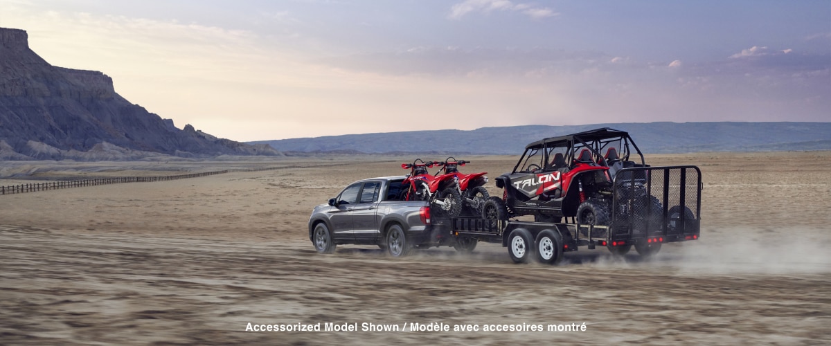 Rear side wide view of grey Ridgeline driving in the desert with two motocross bikes in truck-bed, towing a trailer with a dune buggy.
