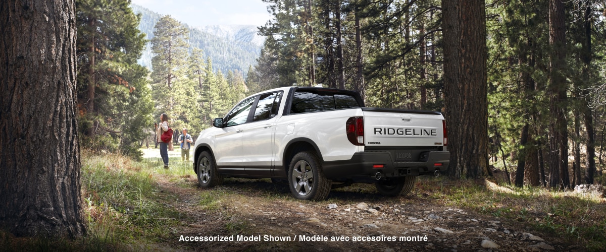 3/4 rear view of white Ridgeline parked in forest. A couple standing in front it take in the scenery.