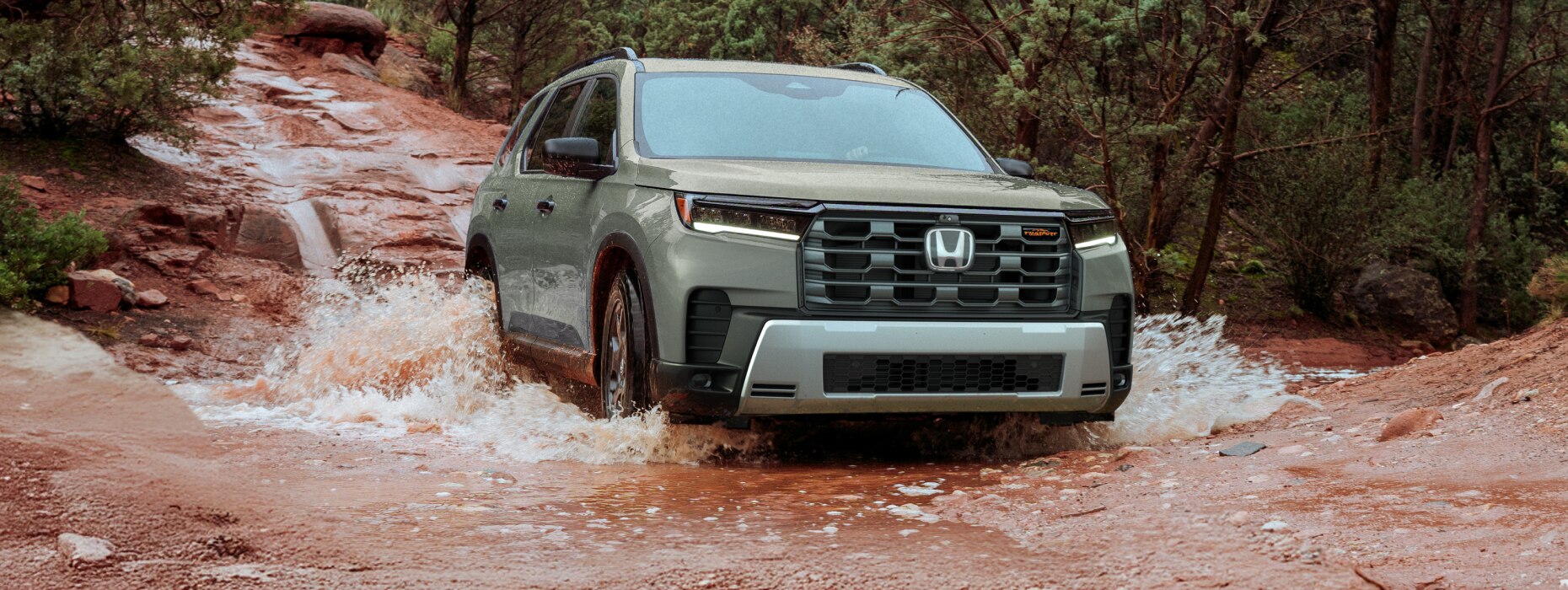 3/4 front wide view of a light green Pilot driving through a muddy creek in a forested area. 