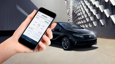 Black Odyssey parked in front of contemporary architecture. Hand in foreground holds a smartphone with the HondaLink™ app. 