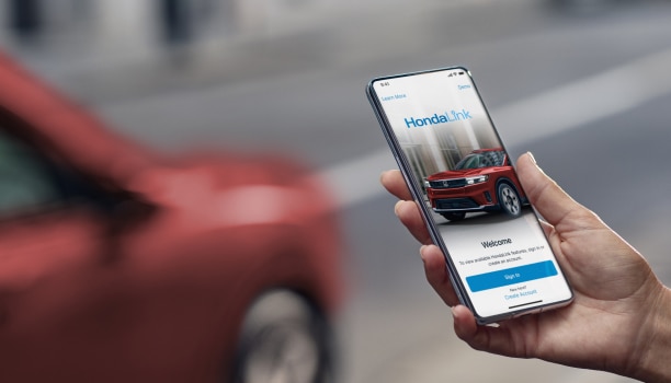 Closeup of a hand holding a smartphone with the HondaLink sign-in page on screen. Behind, out of focus, is a red car.