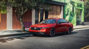 Plan de trois quarts avant d’une Accord rouge stationnée en bordure de rue à côté d’un arbre et devant un bâtiment en briques tendance avec une fenêtre présentant un motif coloré.