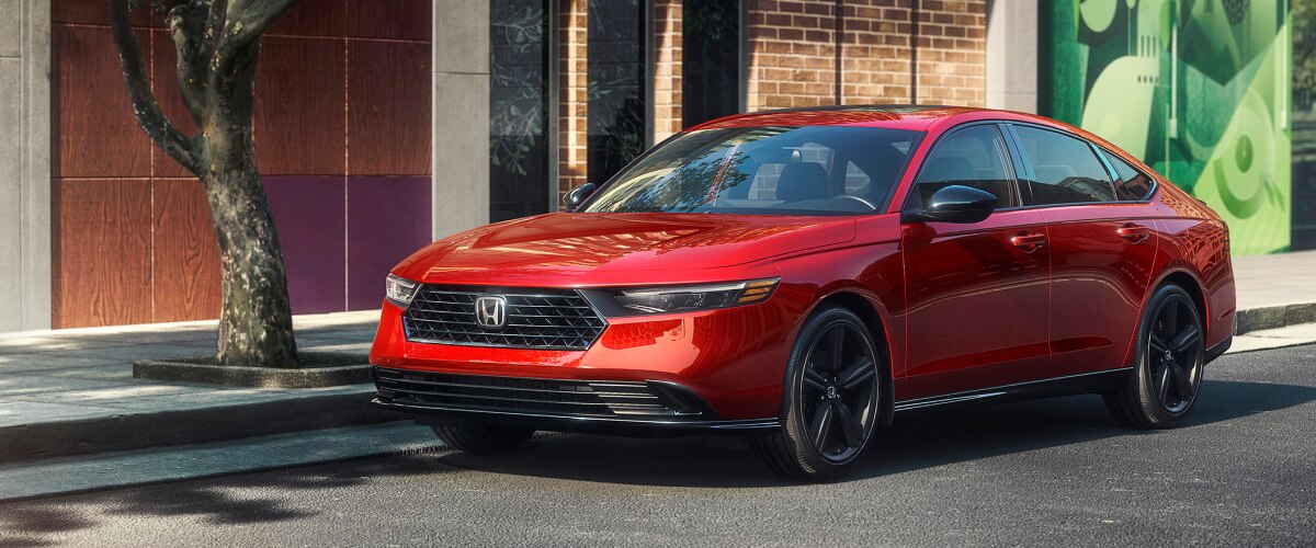 Plan de trois quarts avant d’une Accord rouge stationnée en bordure de rue à côté d’un arbre et devant un bâtiment en briques tendance avec une fenêtre présentant un motif coloré. 