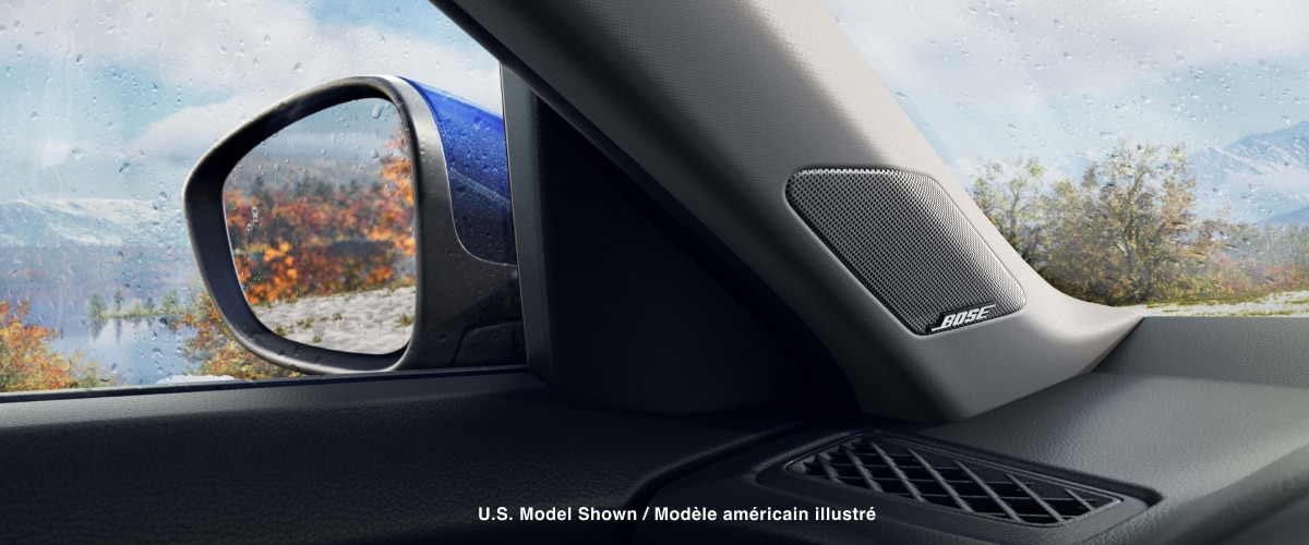 Vue intérieure d’un rétroviseur extérieur et d’un haut-parleur BOSEMD. Vue depuis la vitre du côté conducteur : une chaîne de montagnes, un lac et des arbres aux feuilles orangées.