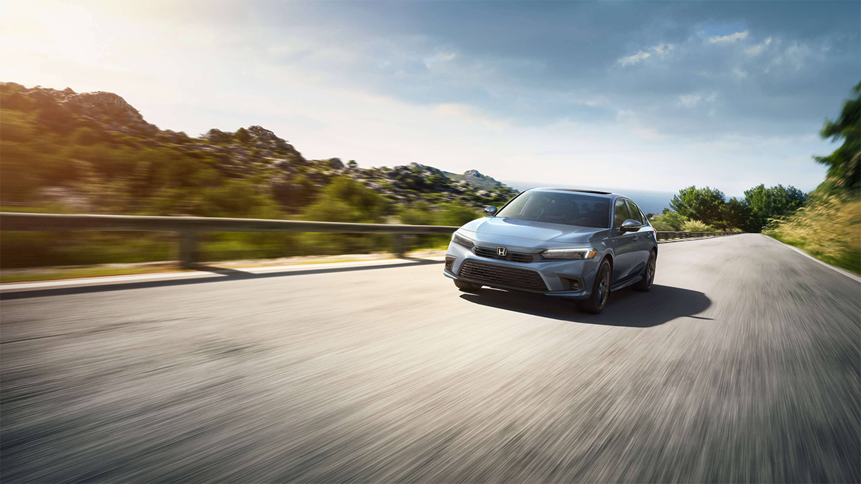 Front view of a grey Honda Civic Sedan driving toward camera on an oceanside highway.