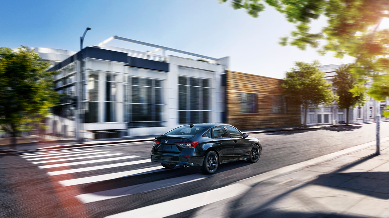 Wide view of a black Civic Sedan driving by upscale boutique architecture on a city street during the day.