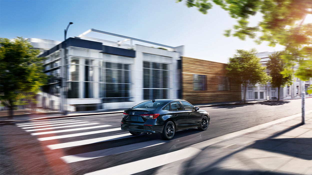 Wide view of a black Honda Civic Sedan driving by upscale boutique architecture on a city street during the day.