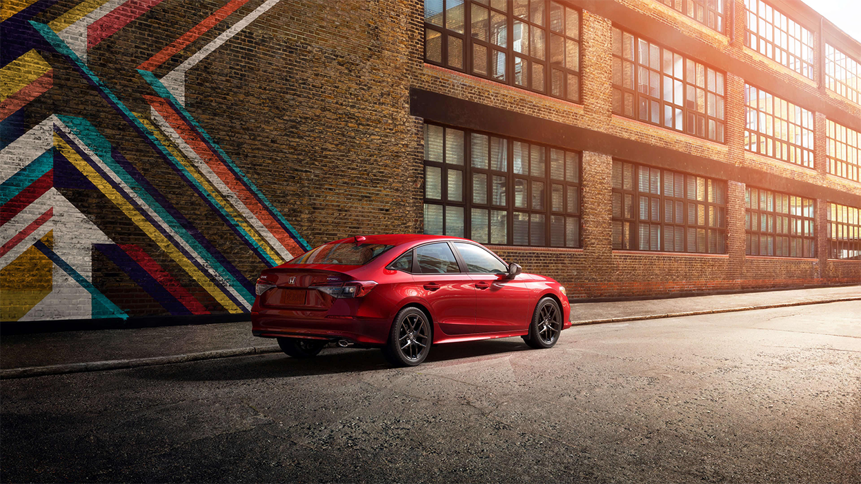 3/4 rear view of red Civic Sedan in an old factory district parked next to a graffitied brick wall.