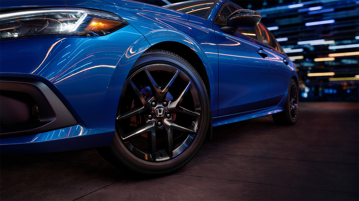 Closeup of the front wheel on a parked blue Civic Sedan in a dark warehouse-like space.