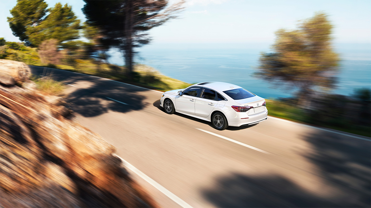 3/4 rear bird’s eye view of white Civic Sedan driving on an oceanside highway in the day. 