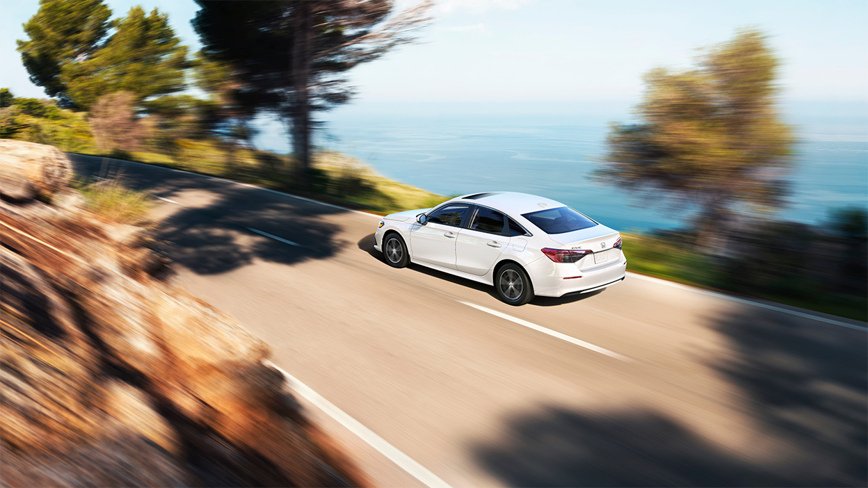 3/4 rear bird’s eye view of white Honda Civic Sedan driving on an oceanside highway in the day. 