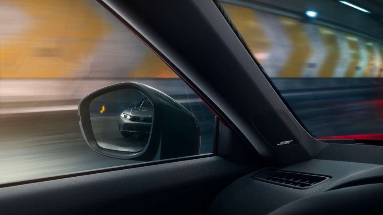 Interior closeup of the door mirror with the reflection of a car in it and a lit up blind spot indicator light.