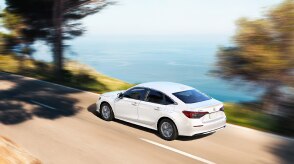 3/4 rear bird’s eye view of white Civic Sedan driving on an oceanside highway in the day.