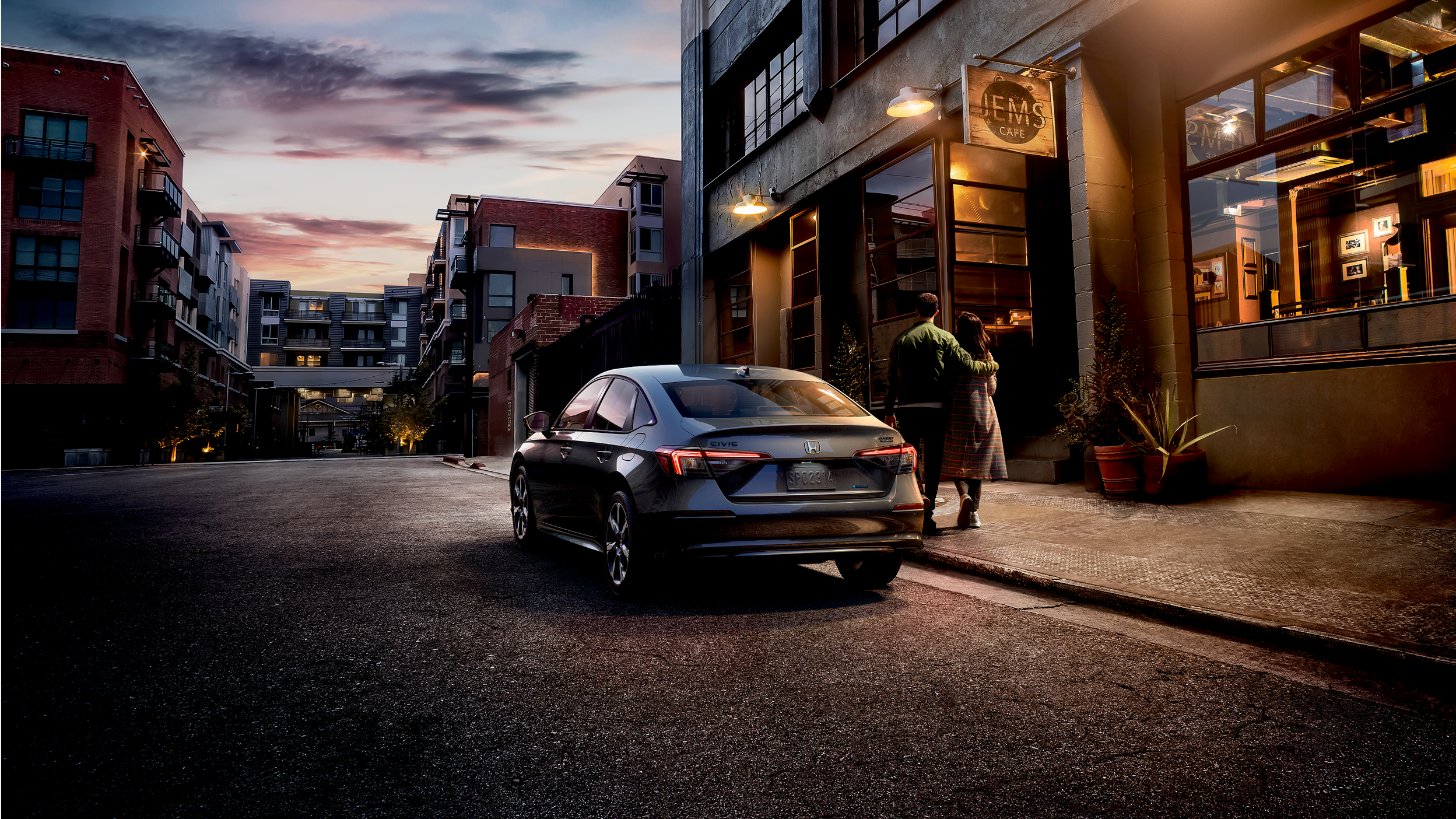 3/4 rear wide view of grey Civic Sedan parked in a trendy city street at dusk. A couple walks toward a cafe, away from the Civic.