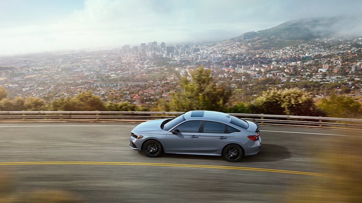 3/4 bird’s eye sideview of a greyish blue Civic Si driving highway overlooking a city.