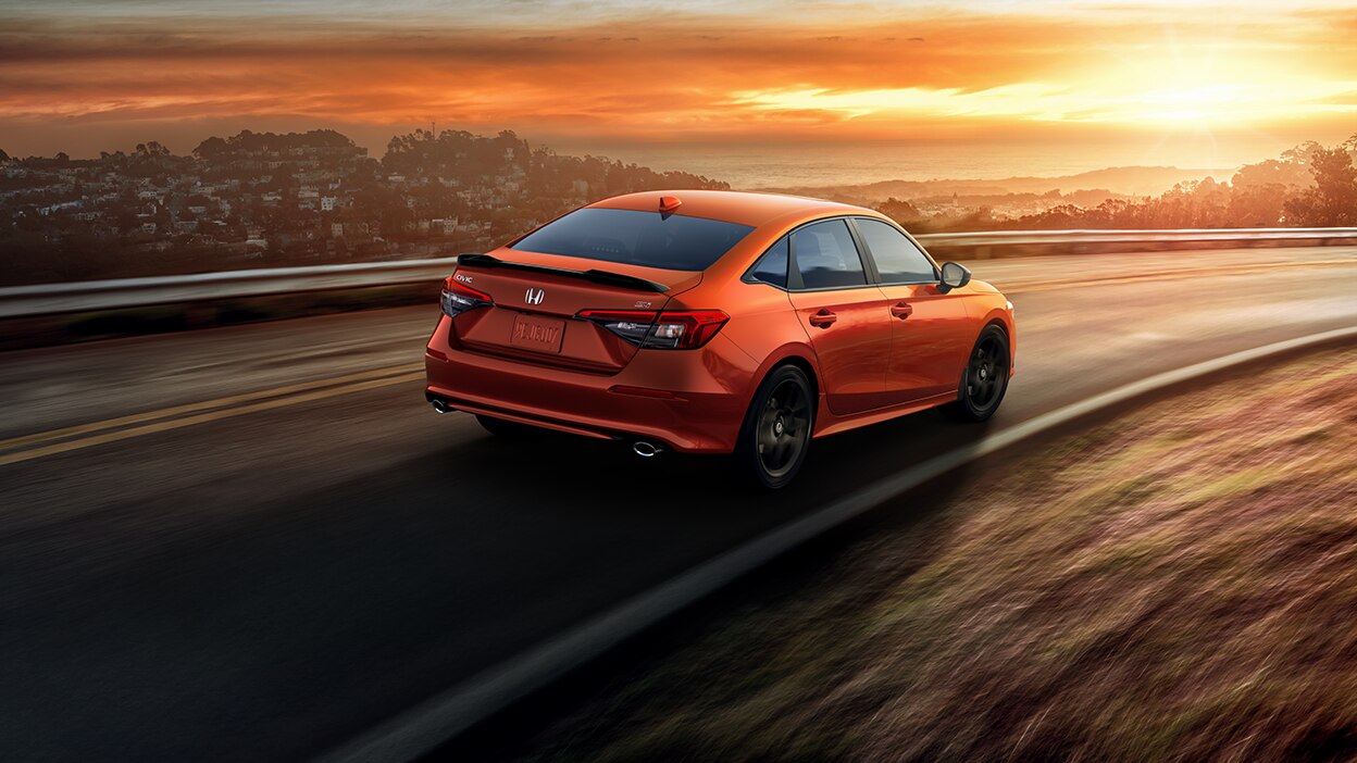 3/4 rear view of orange Civic Si driving on a highway with an ocean view at sunset.