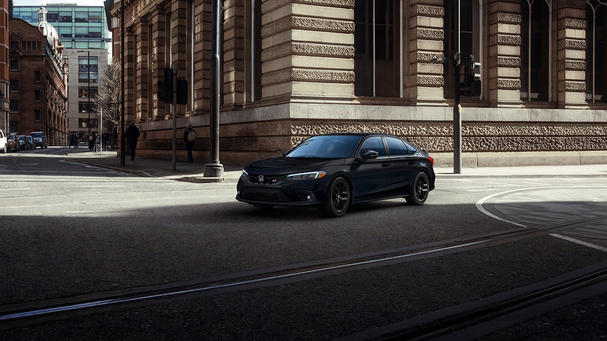 3/4 front sideview of black Civic Si taking a turn on a city street near an old building.