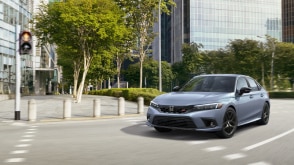 3/4 front view of greyish blue Civic Si taking a turn on a city street with lots of greenery on a sunny day.
