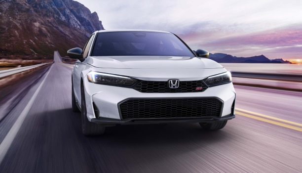 Front view of white Civic Si driving toward camera on a mountainous oceanside highway.
