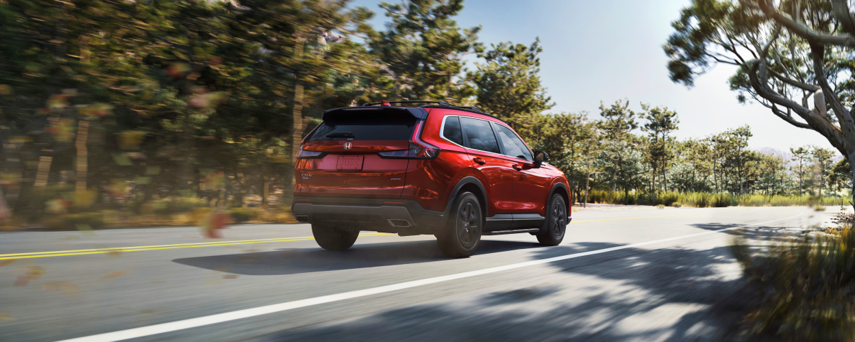 3/4 rear view of red 2023 CR-V driving on a forest highway. 