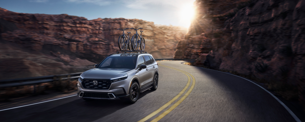 3/4 front view of grey CR-V, with two bikes on the roof racks, driving on a winding desert canyon highway.