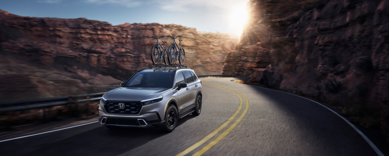 3/4 front view of grey Honda CR-V, with two bikes on the roof racks, driving on a winding desert canyon highway.