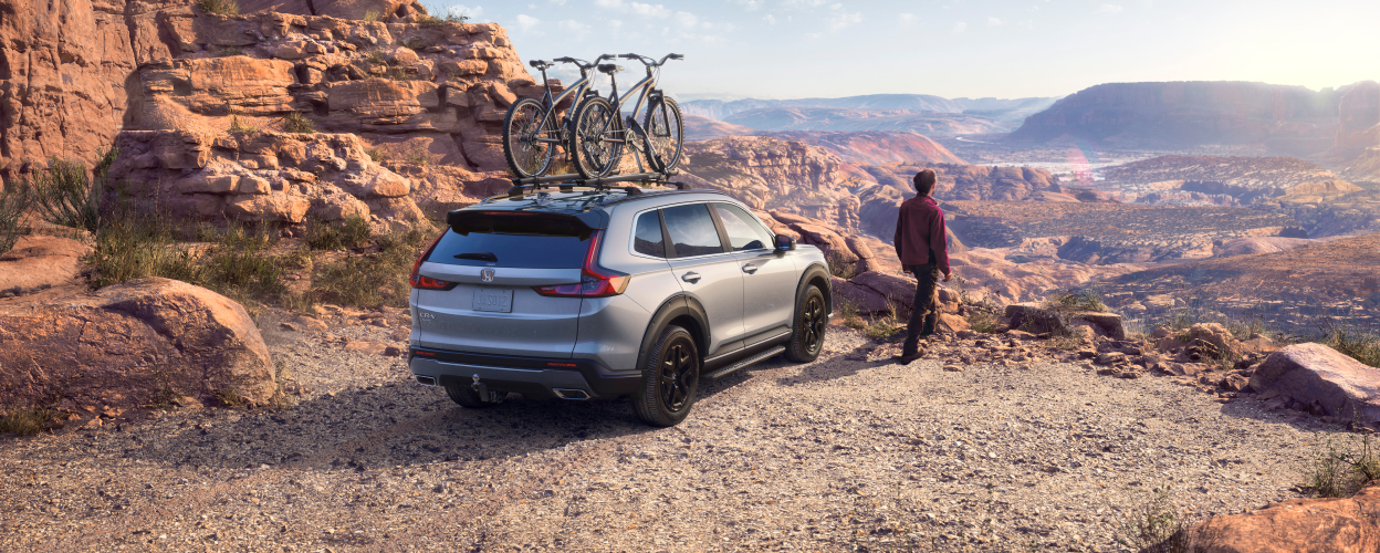 3/4 rearview of grey CR-V with two bikes on the roof racks and a man standing next to it, taking in the desert canyon view.