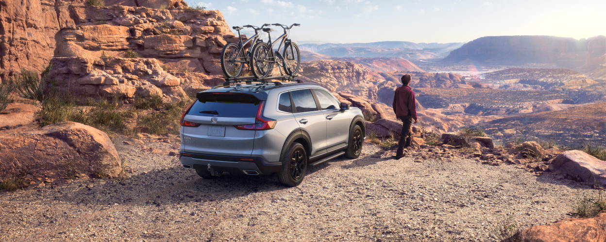 3/4 rearview of grey Honda CR-V with two bikes on the roof racks and a man standing next to it, taking in the desert canyon view.