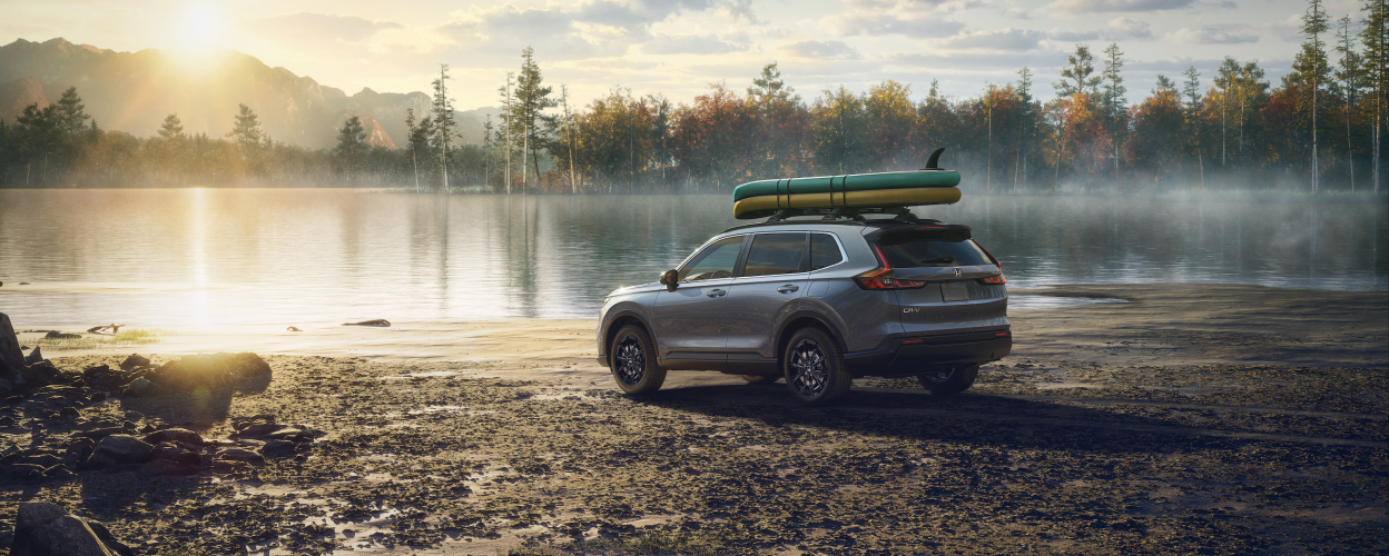 3/4 rear sideview of grey 2023 CR-V, with two paddle boards on the roof racks, parked on a lake shoreline on a dewy morning.