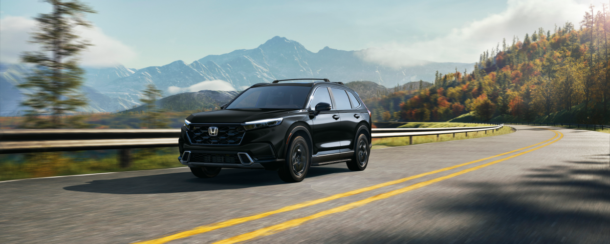3/4 front view of black 2023 CR-V driving on a winding forest highway with fall-coloured trees and mountain range as backdrop.