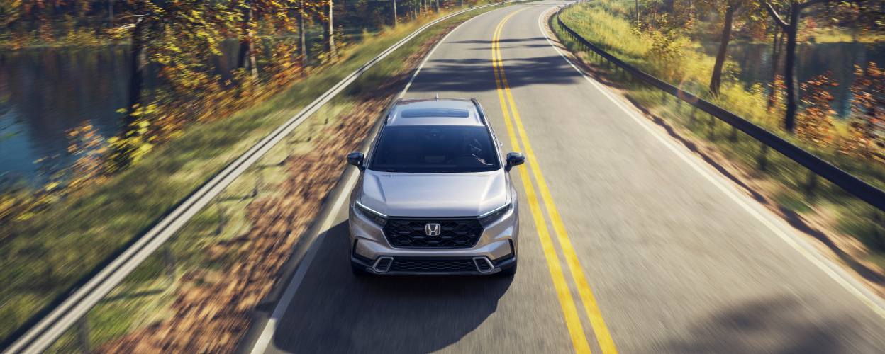 3/4 front bird’s eye view of grey 2023 CR-V driving toward camera on a lakeside highway.