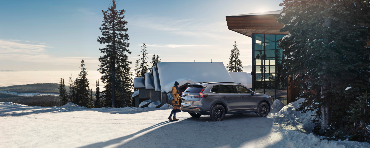Woman in winter attire opening the tailgate of a grey CR-V outside a snowy ski chalet.
