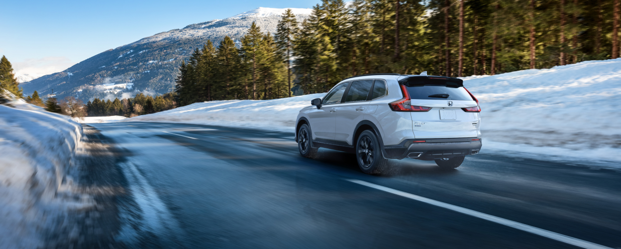 3/4 rear view of white CR-V driving on highway in a snowy forest.