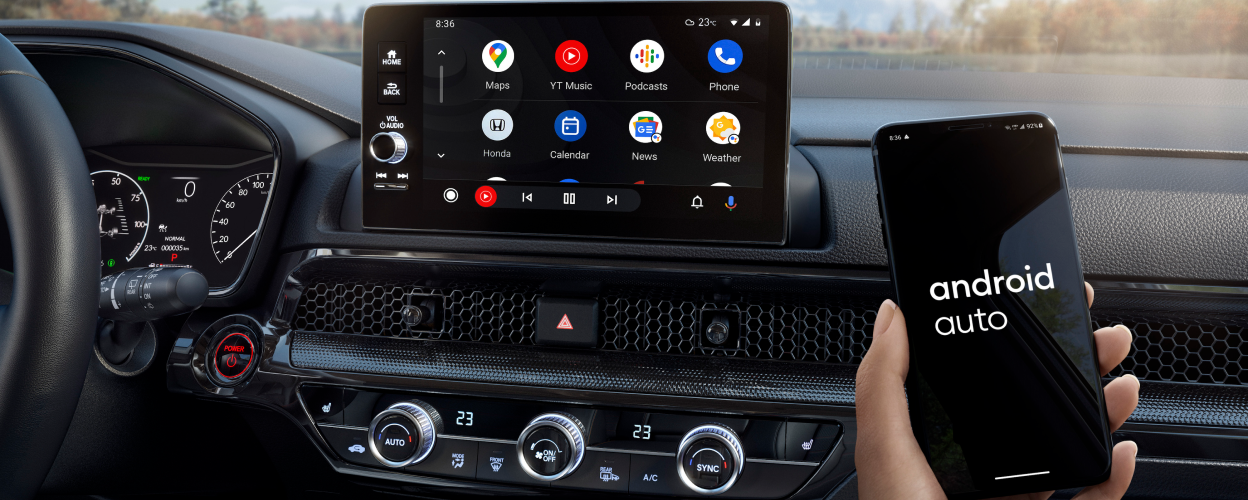 Closeup of touchscreen display. In foreground, a hand holding a smartphone that reads “Android Auto” on screen. Forest backdrop out windshield. 
