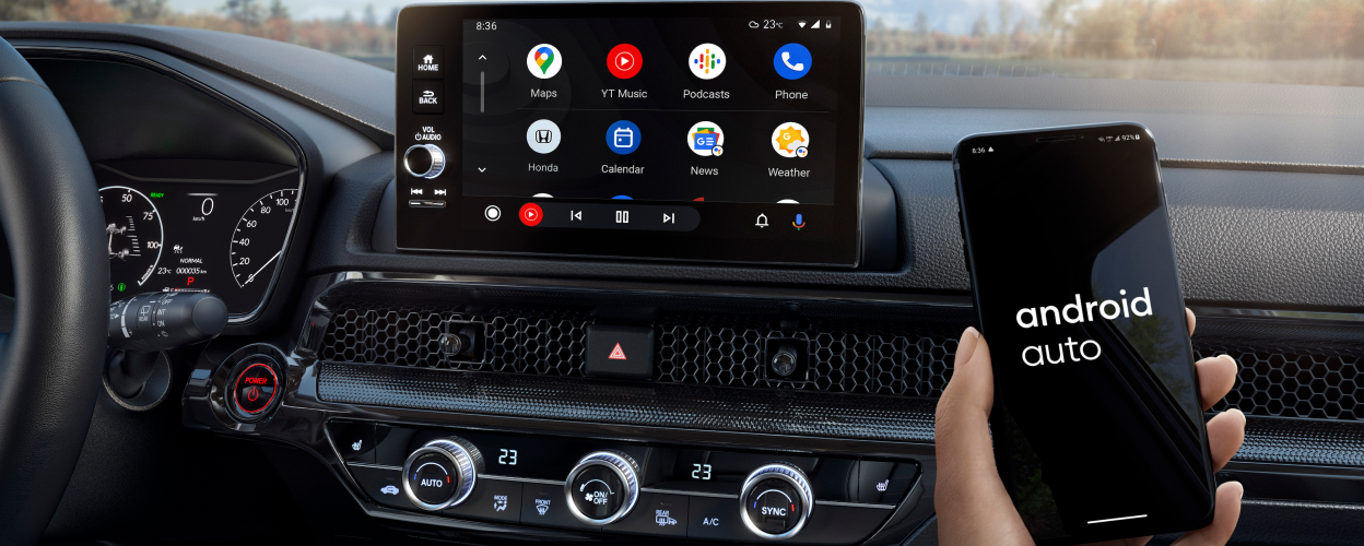 Closeup of touchscreen display. In foreground, a hand holding a smartphone that reads “Android Auto” on screen. Forest backdrop out windshield.