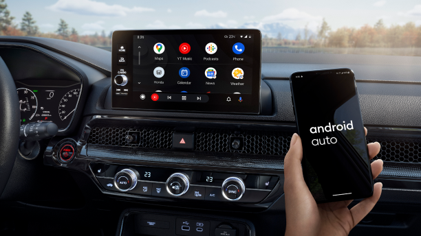 Closeup of touchscreen display. In foreground, a hand holding a smartphone that reads “Android Auto” on screen. Forest backdrop out windshield. 