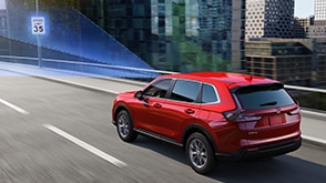 3/4 rear view of red CR-V driving over city bridge. Blue sensor lines emit from the front, detecting the traffic sign ahead.