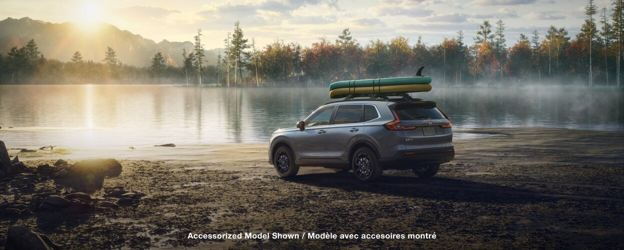 Vue latérale arrière 3/4 d’un CR-V gris, avec deux planches à pagaie sur la galerie de toit, stationné sur le bord d’un lac au petit matin. 