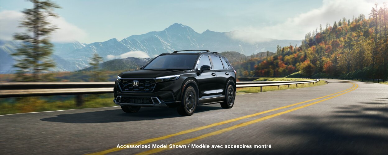 Vue avant 3/4 d’un CR-V noir roulant sur une route sinueuse au milieu d’une forêt avec arbres en automne et chaîne de montagnes en arrière-plan.