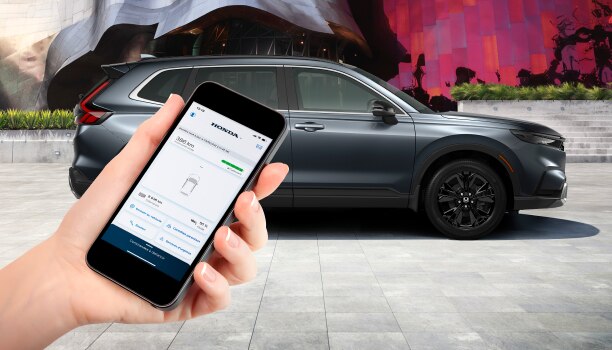 Vue latérale d’un CR-V gris stationné devant un mur avec des graffitis. Une main au premier plan tient un téléphone intelligent affichant l’application HondaLink.