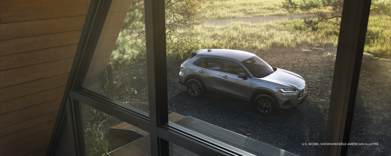 3/4 bird’s eye view of a grey HR-V parked in the countryside, seen through the windows of an a-frame cottage. 