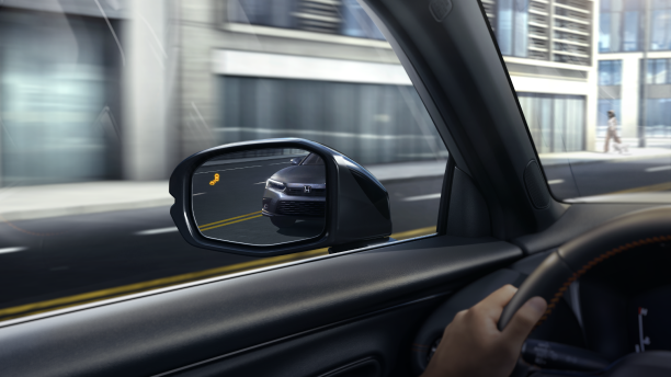 Interior closeup of the door mirror with the reflection of a car in it and a lit up blind spot indicator light. 