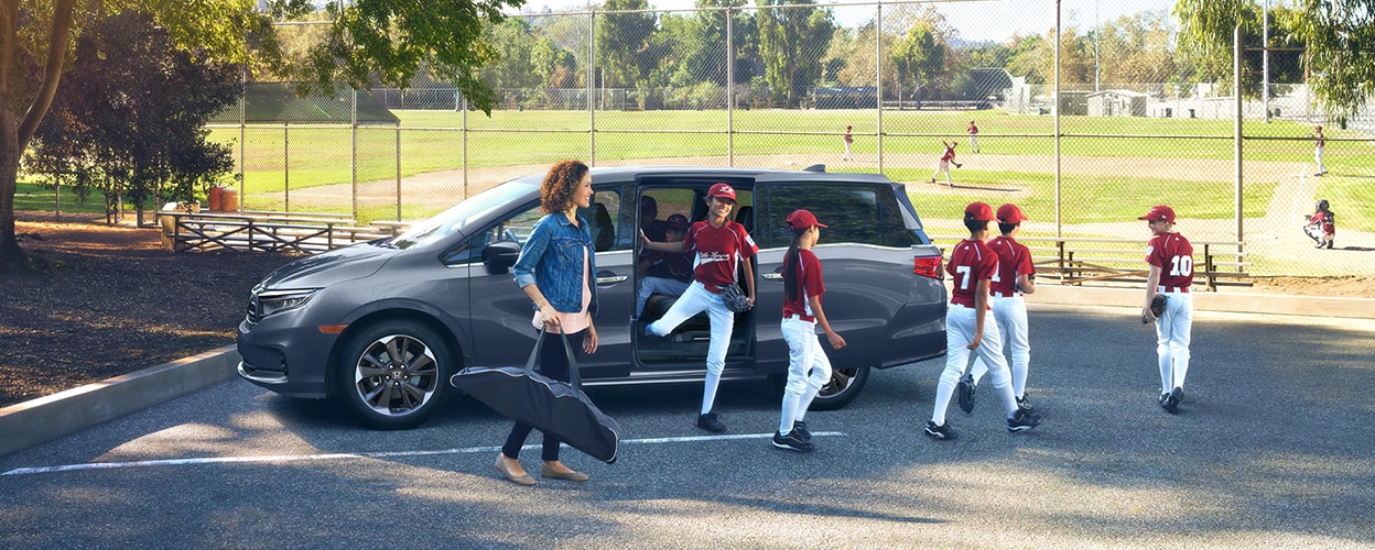 Mom and kids’ baseball team exiting a grey Honda Odyssey parked near a baseball diamond. 