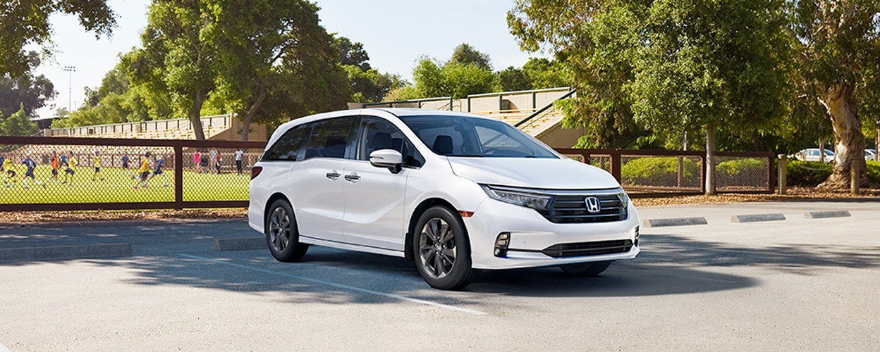 3/4 front view of white Honda Odyssey parked near a soccer field. 