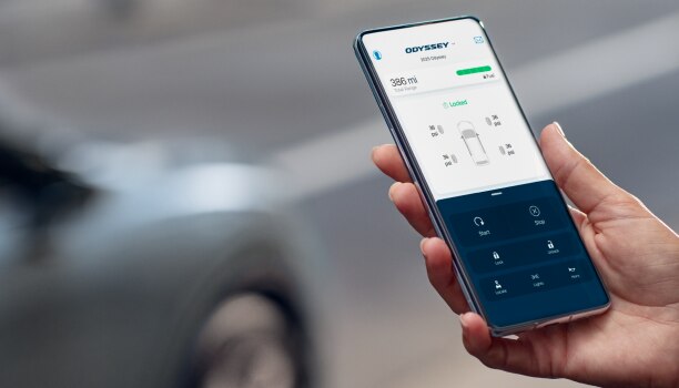 Mains d’une personne qui utilise l’application HondaLink sur son téléphone. Odyssey grise stationnée en arrière-plan.  