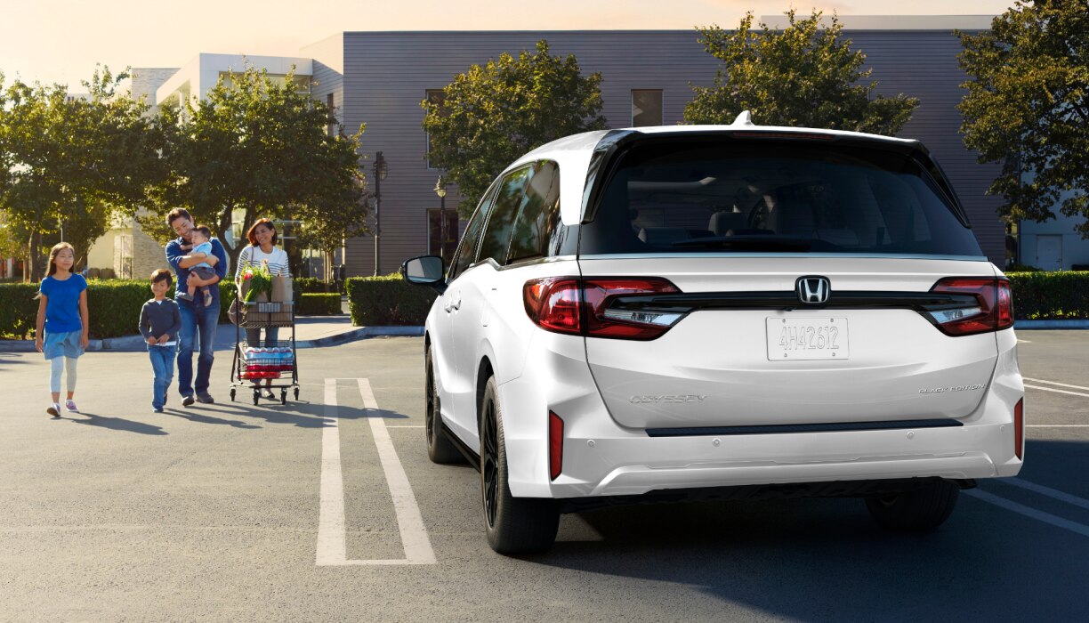 Vue arrière d’une Odyssey grise dans un stationnement vide. Une famille marche vers le véhicule, en poussant un panier d’épicerie.