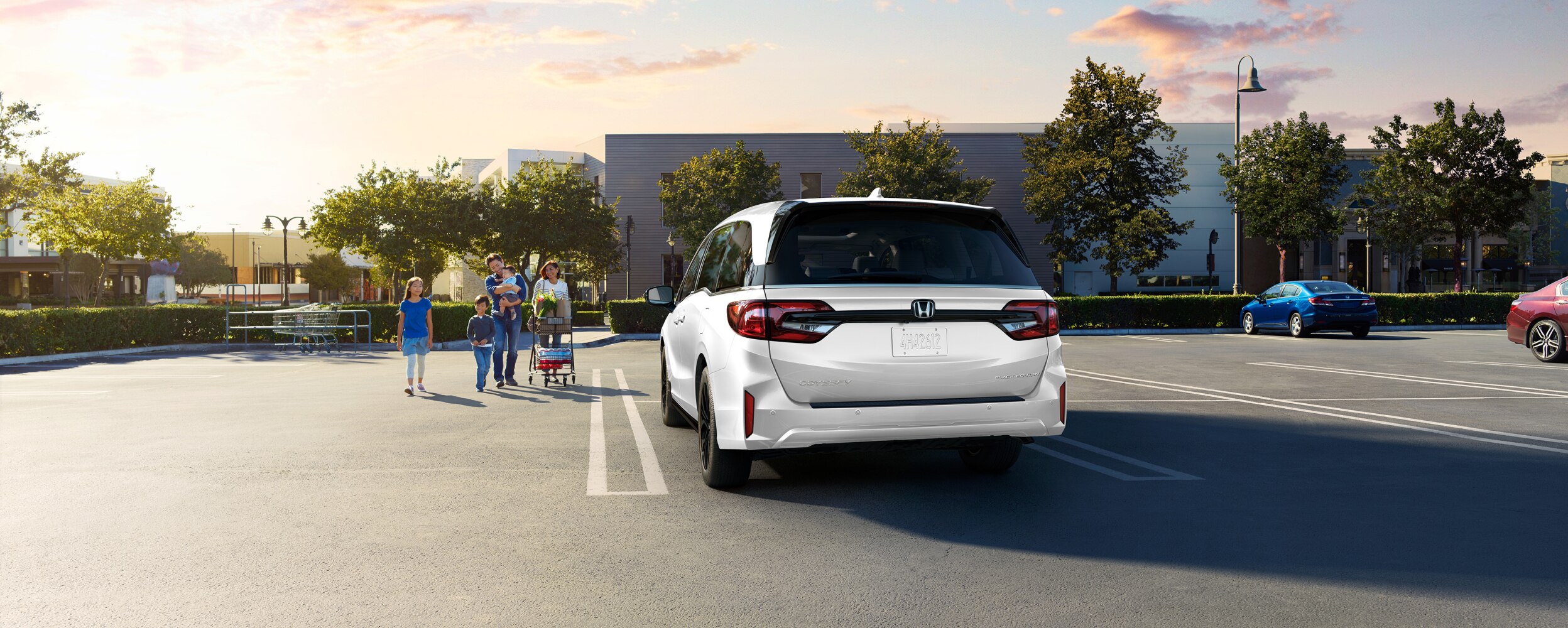Vue arrière d’une Odyssey grise dans un stationnement vide. Une famille marche vers le véhicule, en poussant un panier d’épicerie.