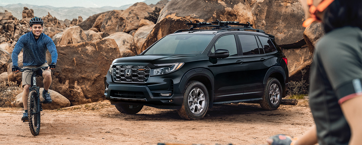 3/4 side front view of black Honda Passport parked in desert; two mountain biking friends near it.