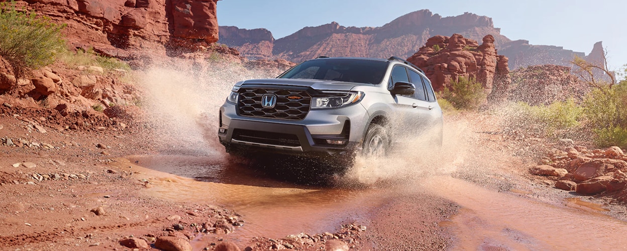 3/4 front view of grey Honda Passport plowing through water on desert trail.