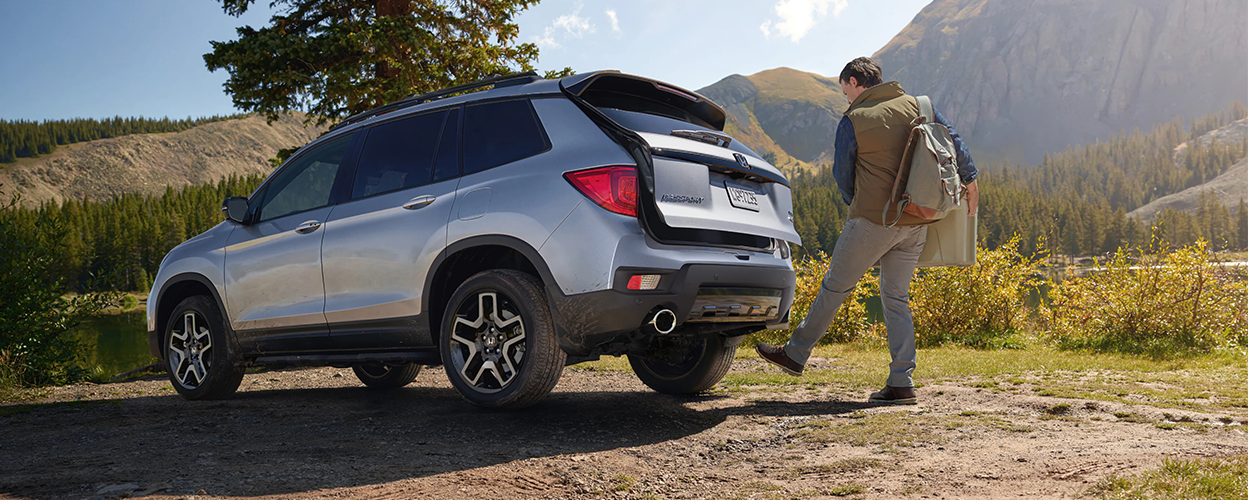3/4 rear view of grey Honda Passport, outdoors. Man waving foot under bumper, opening trunk.